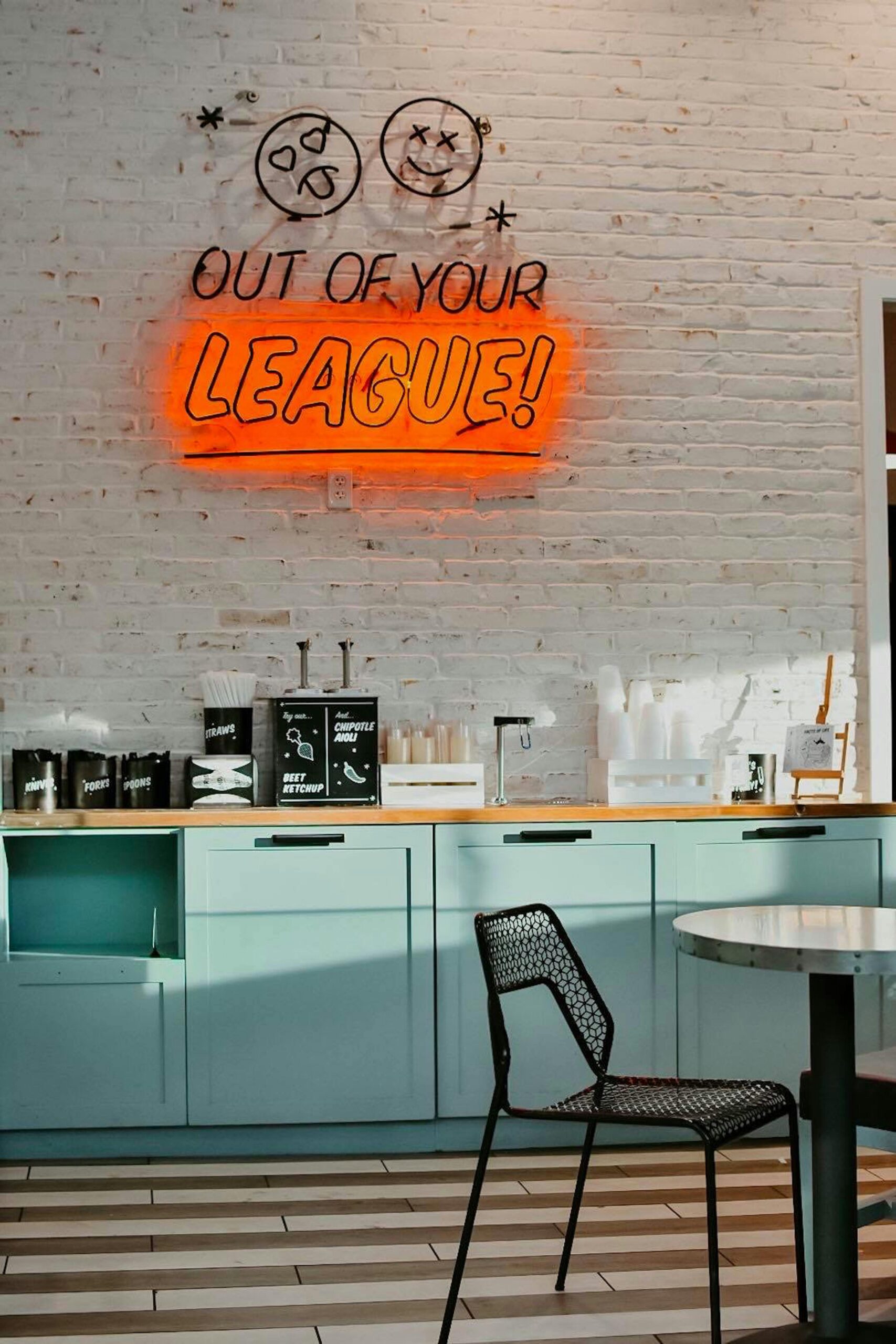 Trendy cafe-style interior with mint green cabinetry, white brick wall, black mesh chair, and bold neon sign reading “Out of Your League!”, designed by SKDesignz.