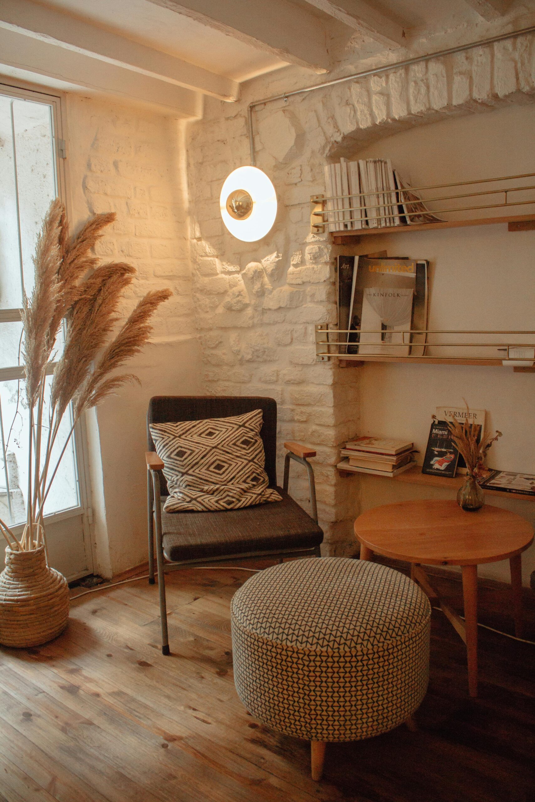 Warm boho-style reading nook with textured white brick walls, woven pampas grass decor, modern accent chair, patterned ottoman, and wood flooring, designed by SKDesignz interior studio.